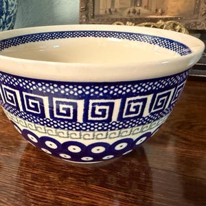 May include: A ceramic bowl featuring a cream-coloured interior and a blue and white exterior. The exterior pattern includes a band of geometric Greek key motifs, a dotted border, and a lower section decorated with repeating blue circular eye designs.