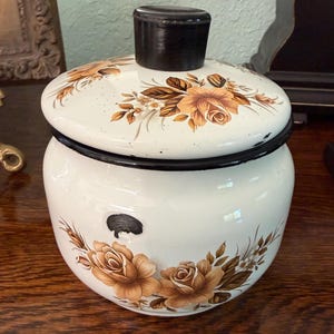 May include: A cream-colored enamel canister with a matching lid and a dark brown knob handle. The canister features a floral pattern of brown roses and green leaves on both the body and the lid, with a black rim and a small black circular mark.