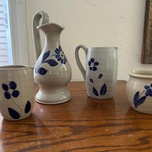 May include: A collection of handmade pottery with a floral design. The set includes a pitcher, two mugs, and a small bowl. Each piece is off-white with blue floral accents. The pottery is displayed on a wooden surface.
