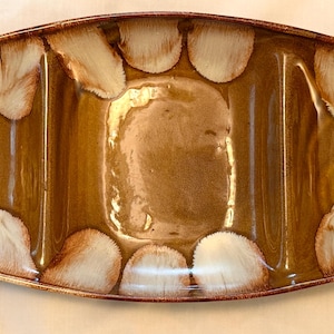 May include: An elongated, oval-shaped ceramic serving dish with three distinct compartments. The dish features a glossy, warm brown glaze with a decorative border of white, petal-like patterns radiating inward along the curved edges.