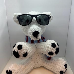 May include: A handmade white crochet polar bear wearing black sunglasses and a blue and burgundy scarf. The bear has black paws and a black nose. The bear is sitting with its arms crossed.