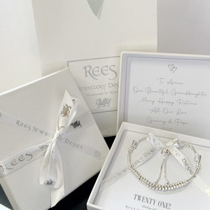 May include: A white gift box with a silver ribbon and a silver dragon design. The box is open and contains a silver bracelet with a charm. The bracelet is on a white background with the text "Twenty One!" and "Rees Jewellery Design Handmade in Wales".