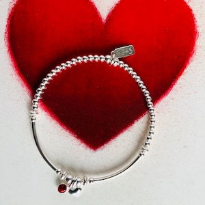 May include: A silver beaded bracelet with a red gemstone and a heart charm. The bracelet is set against a red heart shape on a white background. A small tag with the word "KEES" is attached to the bracelet.