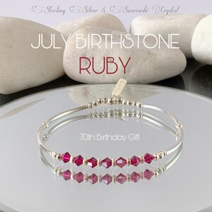 May include: A silver bracelet with a row of red Swarovski crystals. The bracelet is on a reflective surface and has a tag that says "Rees". The text "70th Birthday Gift" is also visible.
