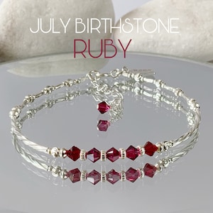 May include: A silver bracelet featuring a row of red ruby crystals. The bracelet has a delicate, twisted design with a small charm hanging from it. The text "JULY BIRTHSTONE RUBY" is displayed above the bracelet.