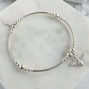 May include: A silver bangle bracelet with a small bee charm. The bracelet is made of silver beads and has a delicate design.