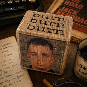 May include: A wooden block with the words "burn burn burn" printed on top and a portrait of a man on one side. The block is surrounded by a typewriter, a book titled "On The Road" by Jack Kerouac, a coffee mug, and a notebook with handwritten text.