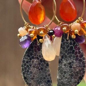 May include: Gold hoop earrings with teardrop-shaped orange gemstones and a cluster of smaller, colorful beads. The earrings also feature a dark, textured, leaf-shaped pendant. The design incorporates amethyst, amber, and other colorful stones.
