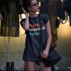 Pu&ograve; includere: Una maglietta grigia scura con il testo "Nasty women vote." stampato in lettere colorate. La maglietta &egrave; indossata da una persona con occhiali e uno chignon.