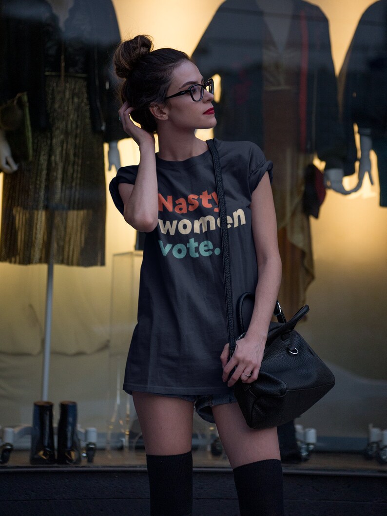 Pu&ograve; includere: Una maglietta grigia scura con il testo "Nasty women vote." stampato in lettere colorate. La maglietta &egrave; indossata da una persona con occhiali e uno chignon.