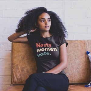 Pu&ograve; includere: Una maglietta nera con il testo "Nasty women vote." in un font in stile retr&ograve; con colori rosso, arancione, giallo e verde.