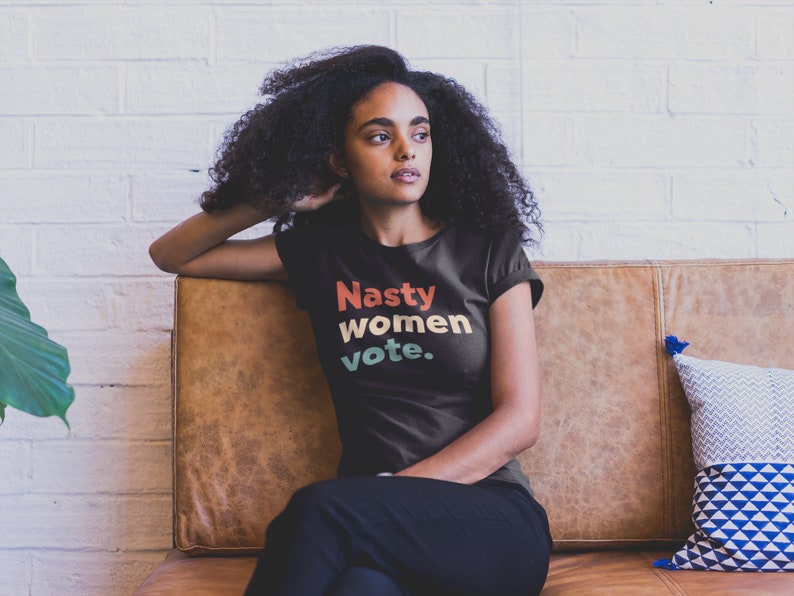 Pu&ograve; includere: Una maglietta nera con il testo "Nasty women vote." in un font in stile retr&ograve; con colori rosso, arancione, giallo e verde.