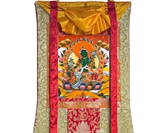 Può includere: Una divinità verde con una corona e un fiore di loto siede su un trono con uno sfondo giallo. La divinità è circondata da un'aureola e da un mandala. L'immagine è incorniciata da un bordo rosso e oro ed è montata su un rotolo.