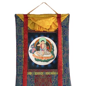 May include: A colorful, rectangular wall hanging featuring a central image of a seated figure within a white circle, surrounded by a blue background with stars. The hanging has a patterned blue and red fabric with red ribbons and a yellow top.