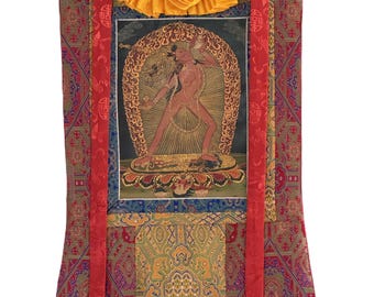 May include: A Tibetan thangka painting featuring a central figure in a dynamic pose, surrounded by an ornate golden halo. The artwork is set against a dark background and framed by colorful, patterned fabric with red ribbons and a wooden dowel at the bottom.