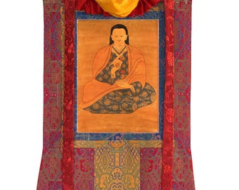 Può includere: Un arazzo verticale da parete con un dipinto centrale di una figura seduta in vesti arancioni e scure a motivi. Il dipinto è incorniciato da bordi in tessuto rosso, blu e oro con intricati motivi intrecciati e due lunghi nastri rossi.