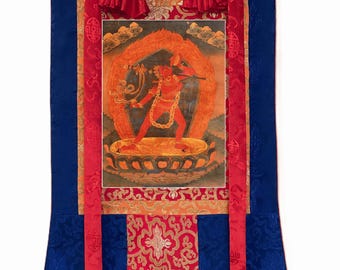 May include: A vertical wall hanging scroll featuring a central painting of a red deity surrounded by flames, standing on a lotus base. The artwork is framed by blue, red, and gold patterned silk fabric borders with two long red ribbons hanging down.