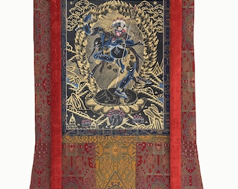 May include: A vertical thangka scroll painting featuring a central dark blue deity figure with multiple arms, surrounded by golden flames. The artwork is framed by a red and gold patterned fabric border with yellow silk accents and a wooden dowel at the base.
