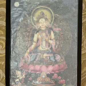May include: A colorful painting of a deity sitting on a lotus flower. The deity is wearing a crown and jewelry and has a serene expression. The painting is framed in black.