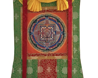 May include: An elaborate wall hanging with a central mandala design. The artwork is framed by green and red fabric with gold accents and a yellow top. The mandala features a complex geometric pattern with vibrant colours, including orange, blue, and red.