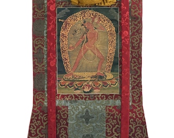 May include: A vertical wall hanging scroll featuring a central painting of a red deity figure surrounded by a golden flame aura. The artwork is framed by red and light blue patterned silk brocade borders, with a gold fabric top and two long red ribbons.