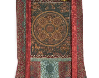 Può includere: Un arazzo verticale da parete con un dipinto mandala circolare centrale nei toni dell'arancione e del marrone. L'opera è incorniciata da bordi in seta a motivi rossi, blu e dorati, con due lunghi nastri rossi che pendono dalla parte superiore.