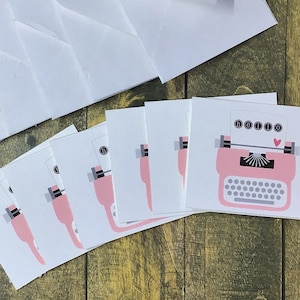 May include: A collection of white envelopes and greeting cards with a pink typewriter design. The cards have a retro aesthetic, with a heart symbol above the typewriter. The background is a wooden surface.