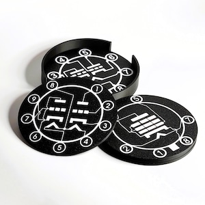 May include: Black round coasters with a white graphic design and numbers 1-9 around the edge. The coasters are in a black holder. The design on the coasters is a series of lines and shapes.