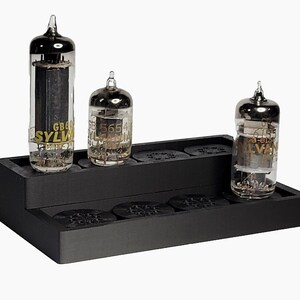 May include: Three vintage vacuum tubes with metallic tops and glass bodies, displayed on a black stepped platform. The tubes have visible internal components and text, including "SYLVANIA" and model numbers. The platform has circular cutouts.