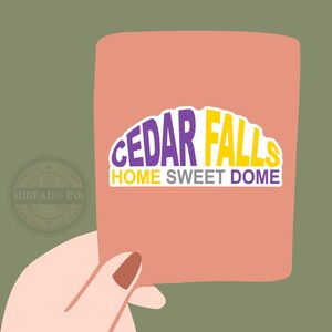 May include: A peach-colored rectangle with the words "CEDAR FALLS HOME SWEET DOME" in purple and yellow. The image is held by a hand with a painted nail. The background is olive green. A circular logo with the text "THREAD & INK CO." is in the lower left corner.