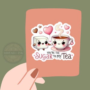 May include: A pink card with a cute illustration of a sugar cube and a teacup holding hands. The sugar cube is holding a pink heart balloon. The text "You're the Sugar to My Tea" is written on the card.
