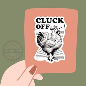 May include: A sticker with a black and white illustration of a rooster with the words "CLUCK OFF" in bold, black letters. The sticker is on a peach-colored background, held by a hand against a green background.