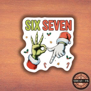 Könnte beinhalten: Ein weißer Aufkleber mit dem Text "SIX SEVEN" in Grün und Rot. Der Aufkleber zeigt eine grüne Hand, die eine "Okay"-Geste macht, und eine weiße Hand, die nach unten zeigt, beide mit Weihnachtsmützen. Zuckerstangen und Sterne sind ebenfalls vorhanden.