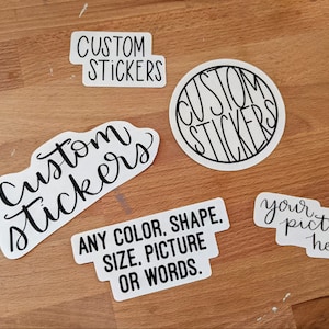 May include: Four white stickers with black lettering. The stickers say "Custom Stickers" in different fonts and styles. One sticker says "Any color, shape, size, picture or words."