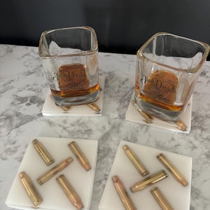 May include: Two white marble coasters with a design of gold bullet casings. Two clear glass shot glasses with Jack Daniels whiskey are sitting on top of the coasters.