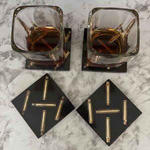 May include: Two square black coasters with a design of gold bullet casings. The coasters are under two square clear glasses filled with amber liquid.