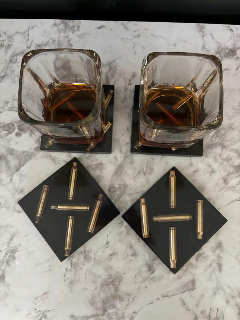 May include: Two square black coasters with a design of gold bullet casings. The coasters are under two square clear glasses filled with amber liquid.