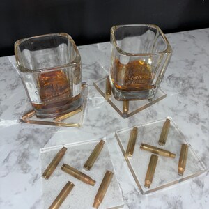 May include: Two clear glass shot glasses filled with amber liquid on bullet-shaped coasters. The coasters are made of clear resin with embedded bullet casings.