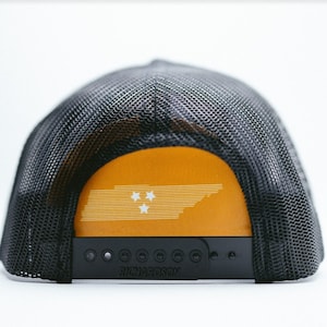 Può includere: Cappellino da camionista nero e arancione con un contorno bianco dello stato del Tennessee e tre stelle bianche. Il cappello ha una rete nera sul retro e le parole "Richardson" stampate sul retro.