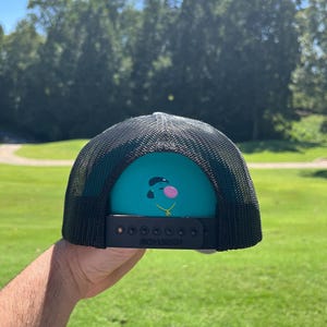 May include: Black trucker hat with a teal front panel featuring a cartoon character with a bubblegum bubble and gold chain. The hat has a black mesh back and a snapback closure.