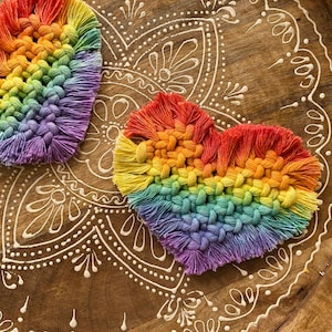 May include: Two rainbow-colored crocheted hearts on a wooden surface with a white mandala design. The hearts are made of yarn in shades of red, orange, yellow, green, blue, indigo, and violet.