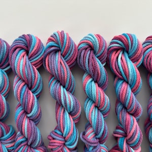 May include: Six skeins of colorful yarn in shades of blue, pink, and purple. The yarn is twisted into a rope-like shape, with each strand showing a gradient of colors. The yarn is likely for knitting or crocheting projects.
