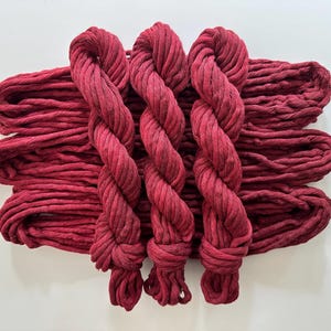 May include: Three deep burgundy yarn skeins arranged vertically over a horizontal base of three additional skeins of the same dark red color. The yarn has a thick, twisted texture and a matte finish, creating a uniform monochromatic appearance.