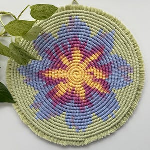 May include: A circular macrame wall hanging featuring a central sunburst pattern in yellow, orange, and magenta, surrounded by light blue petals on a sage green background. The piece is finished with a short, uniform fringe around the edge.