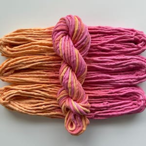 May include: Overhead shot of colorful yarn skeins. Three skeins of orange yarn are on the left, with three skeins of pink yarn on the right. A twisted skein of pink and orange yarn is in the center. The yarn is thick and appears to be cotton.