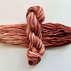 May include: Two skeins of chunky yarn in shades of red, brown, and cream. One skein is a twisted bundle, and the other is a loose, elongated pile. The yarn is thick and appears soft, suitable for various crafting projects.
