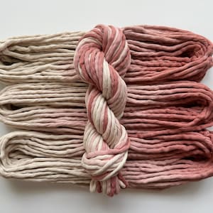May include: Three skeins of thick cotton cord arranged horizontally, featuring an ombre color transition from cream on the left to dusty rose on the right. A central braided knot binds the three skeins together, showcasing the blended color gradient.