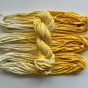 May include: Three skeins of yellow and white yarn are arranged next to a twisted skein of yellow and white yarn. The yarn is a bright yellow color, with white accents. The yarn is likely used for knitting or crocheting.