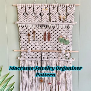 Könnte beinhalten: Cremefarbener Makramee-Schmuck-Organizer mit Holzstäben und Fransen. Der Organizer zeigt verschiedene Ohrringe und Halsketten. Der Text unten lautet "Macrame Jewelry Organizer Pattern".