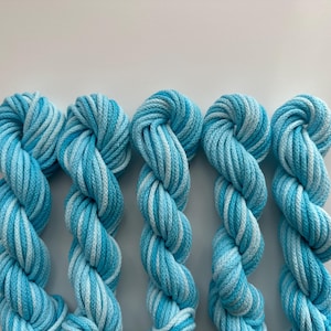 May include: Five skeins of blue and white variegated yarn are arranged in a row. The yarn is twisted into a rope-like shape, with varying shades of light and dark blue and white. The yarn is likely for knitting or crochet projects.